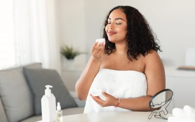 Beauty, cosmetics, skin care concept. Happy black plus size woman holding and smelling cream jar, sitting in bedroom wrapped in towel, free space